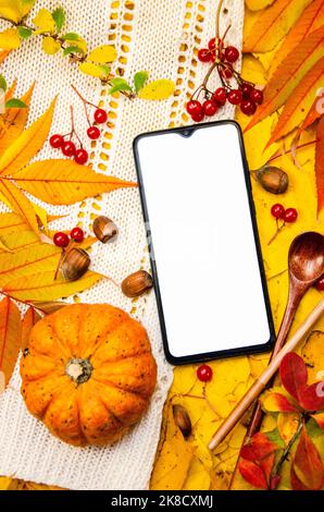 Mockup di uno smartphone display e zucca su uno sfondo in giallo arancione toni rossi: Un bianco lavorato a maglia foglie bacche cucchiai di nocciole. Modello Foto Stock