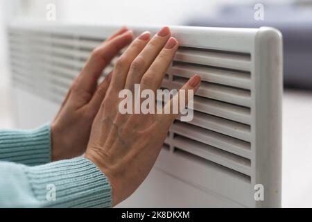 Primo piano di una donna in maglione riscalda le mani sopra la batteria su sfondo di parete bianca. Macro foto delle mani della donna, copia spazio. Concetto di Foto Stock