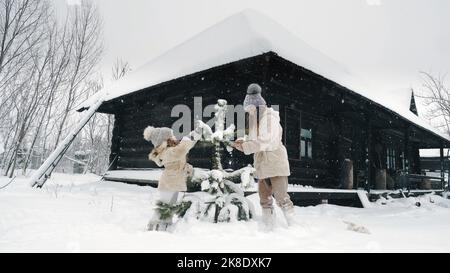 Decorazione esterna dell'albero di Natale. cutie, ragazze graziose decorano il piccolo albero di Natale innevato con i giocattoli di ghiaccio fatti in casa, durante la caduta di neve, nella foresta, vicino alla vecchia casa di legno. Buon tempo il giorno di inverno nevoso. Attività di famiglia di inverno all'aperto. Slow motion. Foto di alta qualità Foto Stock