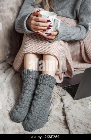 Casa accogliente, donna coperta di coperta calda, bevande caffè e funziona su un computer portatile. Relax, spensierato, stile di vita confortevole. Foto Stock