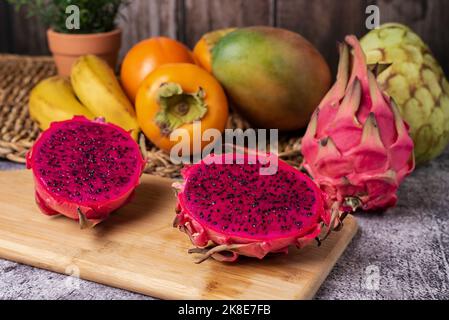 Pitahaya o frutto del drago diviso in due su un asse di legno, esponendo l'interno rosa e i semi neri, sullo sfondo alcuni frutti tropicali come ch Foto Stock