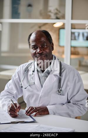 Ritratto del pediatra maschio africano in camice bianco facendo appunti nella scheda paziente a tavola durante il suo lavoro in ufficio Foto Stock