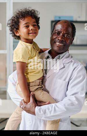 Ritratto di pediatra maschio africano in camice bianco sorridente alla telecamera che tiene piccolo paziente Foto Stock