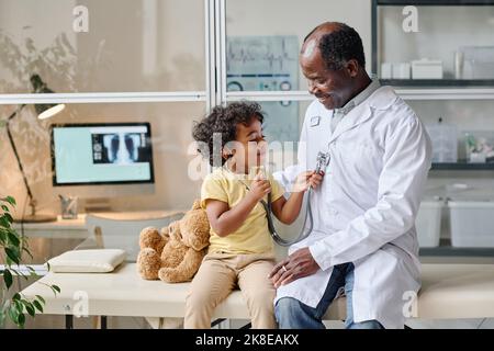 Il ragazzino africano che usa lo stetoscopio per ascoltare il battito cardiaco del medico mentre si siedono sul divano dell'ospedale Foto Stock