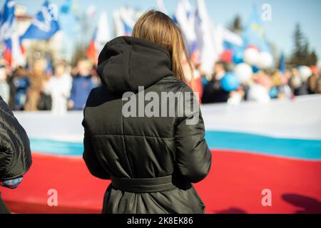 Uomo in raduno in Russia. La ragazza si trova sullo sfondo di persone con bandiere della Russia. Ragazza in giacca nera. Foto Stock