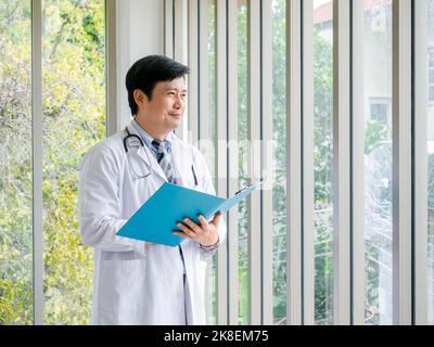 Sorridente uomo asiatico medico ritratto in piedi vicino alle finestre di vetro in ufficio medico con vista verde. Medico asiatico adulto sicuro maschio con stetoscoop Foto Stock