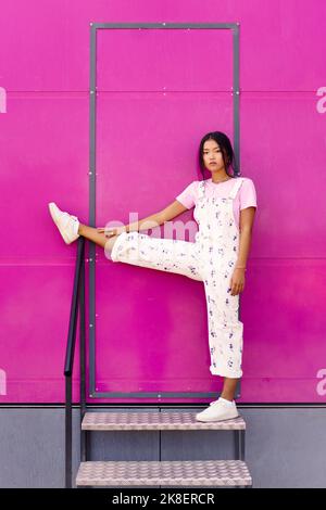 Donna asiatica che guarda la macchina fotografica con espressione seria, sollevando una gamba sopra la ringhiera vicino alla parete rosa. Foto Stock