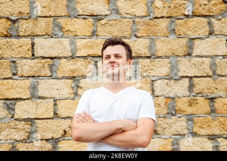 Giovane uomo caucasico di 34 anni in t-shirt bianca con petto a croce su mattoni di pietra calcarea posati su fondo. Stile di vita awa Foto Stock