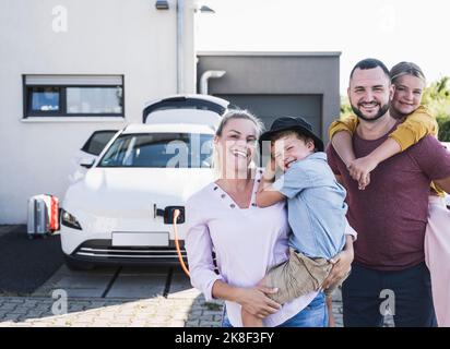 Famiglia felice in piedi di fronte a casa di famiglia e di ricarica auto elettrica Foto Stock