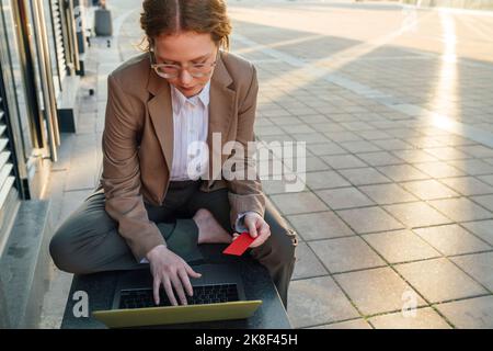 Giovane donna d'affari redhead che paga in linea con la carta di credito usando il laptop Foto Stock