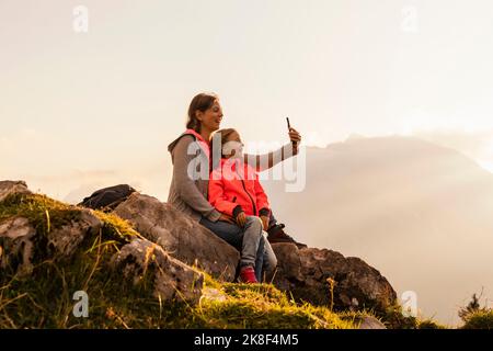 Madre felice con figlia che prende selfie attraverso il telefono cellulare seduto sulla roccia Foto Stock