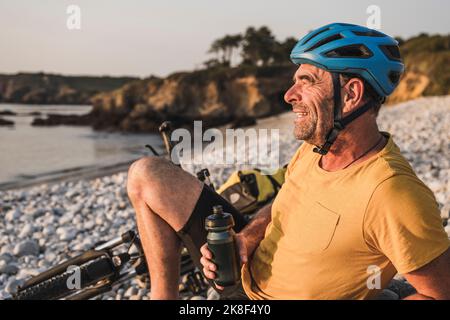 Uomo maturo contemplativo con bottiglia d'acqua che riposa in spiaggia Foto Stock