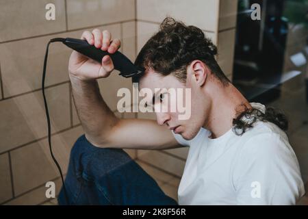 Uomo triste che taglia i propri capelli con rasoio elettrico a casa Foto Stock