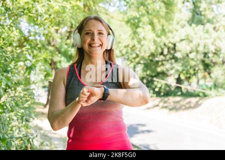 Donna sorridente con cuffie wireless che controllano il polso sul fitness tracker nel parco Foto Stock