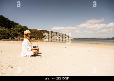 Donna matura che indossa un cappello che pratica la meditazione in spiaggia nelle giornate di sole Foto Stock