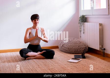 Donna seduta con gli occhi chiusa praticando yoga utilizzando il tablet PC a casa Foto Stock