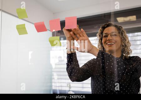 Donna d'affari matura felice che scrive sui biglietti appiccicosi sul vetro all'ufficio Foto Stock