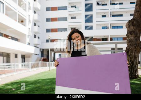 Donna d'affari matura sorridente che tiene il cartello viola bianco in piedi davanti all'edificio degli uffici Foto Stock
