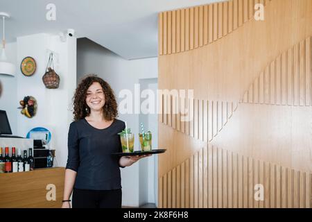 Cameriera giovane e felice con vassoio che serve bevande al ristorante Foto Stock