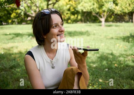 Donna sorridente che invia messaggi vocali tramite smartphone in giardino Foto Stock