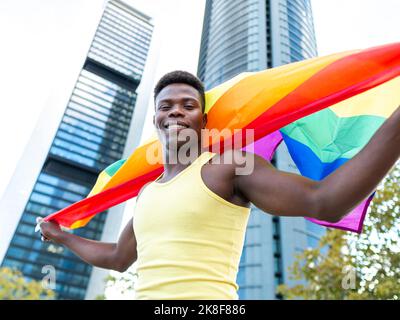 Giovane uomo sorridente che indossa un giubbotto giallo con bandiera arcobaleno davanti agli edifici moderni Foto Stock