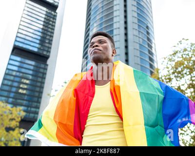 Felice giovane uomo avvolto in bandiera arcobaleno di fronte ai grattacieli Foto Stock