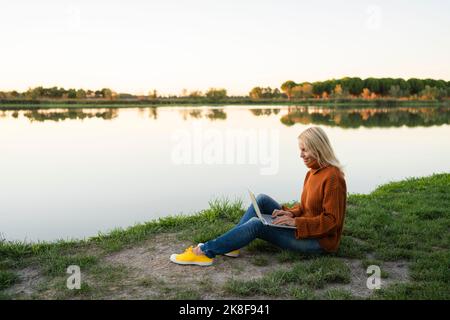 Sorridente freelancer che lavora su un computer portatile seduto vicino al lago Foto Stock