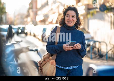 Donna sorridente con smartphone e borsa a piedi in strada Foto Stock