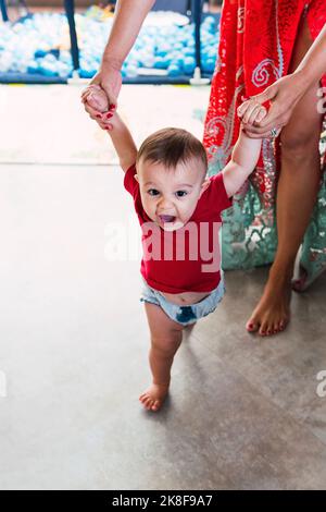 Madre che tiene le mani del bambino che cammina sul pavimento a casa Foto Stock