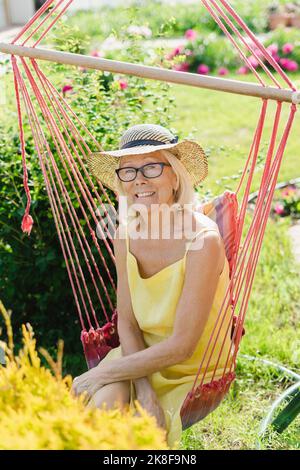 Donna anziana sorridente che indossa un cappello seduto su una sedia sospesa in giardino Foto Stock