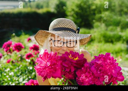 Donna anziana che indossa un cappello che odora fiori rosa freschi in giardino Foto Stock