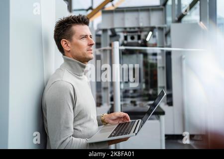 Uomo d'affari maturo e premuroso che tiene il notebook appoggiato al muro nell'industria Foto Stock