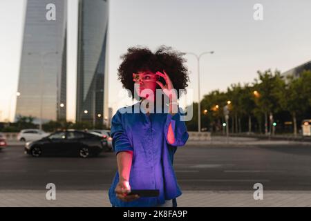 Luce rossa che cade sul viso della donna afro indossando occhiali intelligenti che tengono lo smartphone su strada Foto Stock