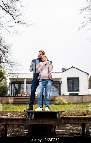 Uomo che abbraccia donna incinta da dietro sul molo di fronte a casa Foto Stock