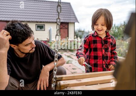 Padre guardando felice figlio pittura swing in legno di fronte a casa Foto Stock