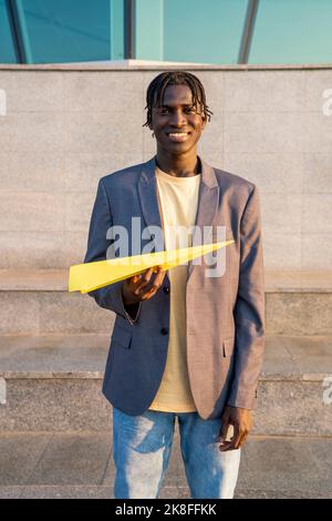 Uomo d'affari sorridente con aeroplano di carta gialla davanti ai gradini Foto Stock