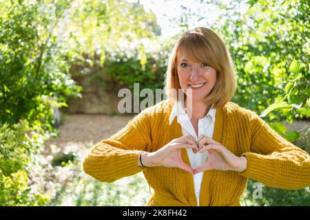 Donna matura sorridente che fa la forma del cuore attraverso la mano in giardino Foto Stock