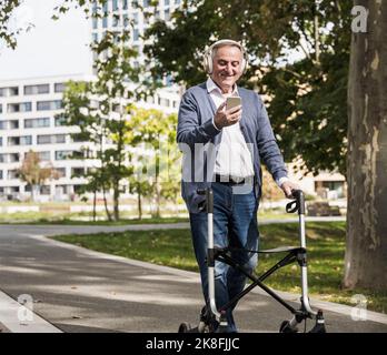 Uomo anziano sorridente che utilizza il telefono cellulare per ascoltare la musica con cuffie wireless che camminano con la mobilità sul sentiero Foto Stock