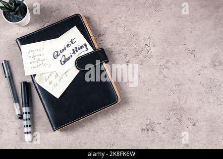 Uscita silenziosa. Vista dall'alto sulla scrivania con articoli da ufficio. Nota adesiva con testo scritto Quiet Quiet Quitting on closed paper business notebook planner. Tranquillo Foto Stock