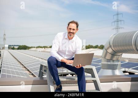 Uomo d'affari maturo seduto con un computer portatile davanti ai pannelli solari Foto Stock