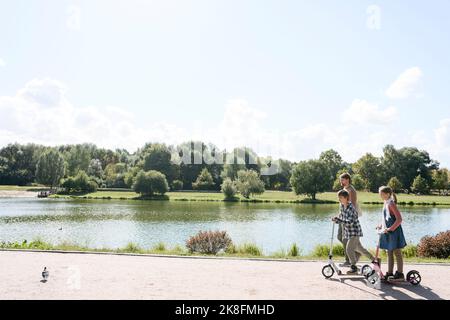 Fratello e sorella a cavallo spingere scooter da padre a piedi nel parco nel fine settimana Foto Stock