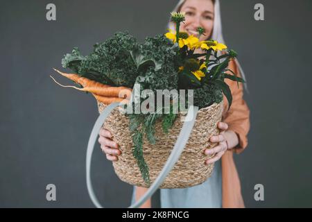 Donna che mostra il sacchetto di vimini riempito di verdure e fiori su sfondo grigio Foto Stock