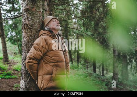 Uomo anziano che indossa una giacca in piedi accanto all'albero nella foresta Foto Stock