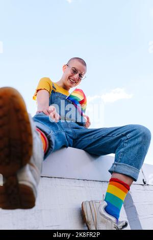 Felice persona non binaria con bandiera arcobaleno seduto sul muro Foto Stock