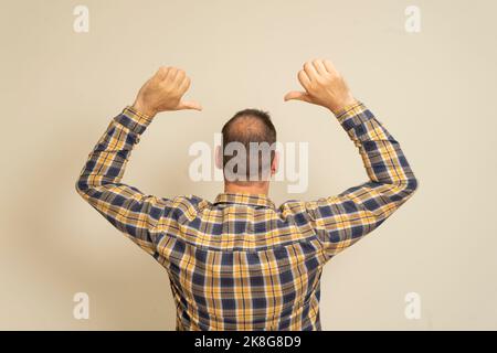 Ritratto di un uomo con incipiente alopecia da dietro, sollevando entrambe le mani e puntando le dita indice verso l'alto, isolato su uno sfondo beige. Foto Stock