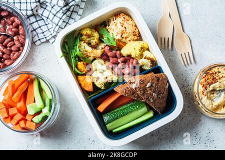 Cottura vegan pranzo scatola piatto. Hummus, fagioli, verdure al forno, insalata verde, pane di segale e bastoncini di verdure in un contenitore di plastica, vista dall'alto. Foto Stock