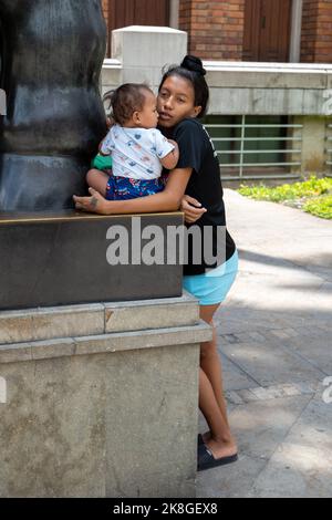 Medellin, Antioquia, Colombia - Settembre 12 2022: Giovane donna dei capelli neri tiene un bambino su una scultura del bronzo, la statua di un grande carattere femminile b Foto Stock