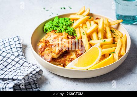 Pesce e patatine fritte. Merluzzo in pastella con patatine fritte e purè di piselli verdi su piatto bianco, fondo grigio. Cucina inglese. Foto Stock