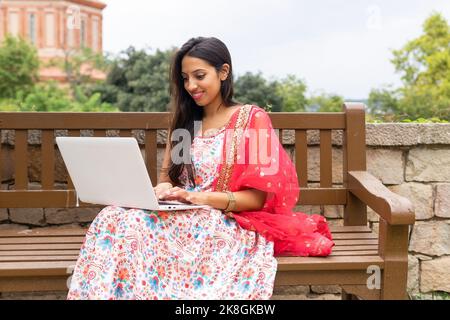 Giovane ragazza indiana freelancer in vestito seduto su una panca di legno e a scrivere sul computer portatile mentre si lavora su un progetto remoto Foto Stock