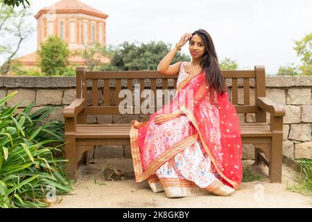Giovane etnia femminile in abito tradizionale seduta su panca di legno nel parco e guardando la macchina fotografica Foto Stock